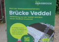 Ein Straßenaufsteller mit einem grünen Plakat, auf dem der Entwurf für eine Brücke zu sehen ist, dazu die Infos