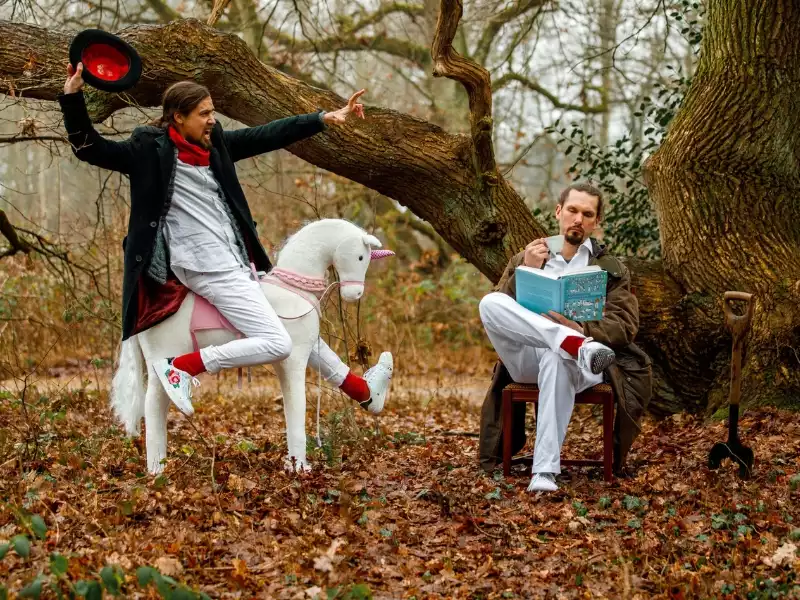 In einem herbstlichen Wald reitet ein Mann auf einem weißen Pfers, ein anderer sitzt und liest ein Buch