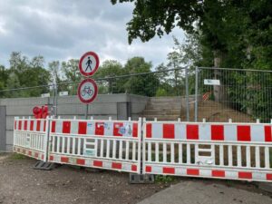 In der unteren Hälfte ein weiß/roter Baustellenabsperrung mit Verbotsschildern für Fußgänger und Radfahrer. Dahinter ein Bauzaun vor einer Betonwand mit einer Treppe an der rechten Seite. Dahinter und rechts Bäume.