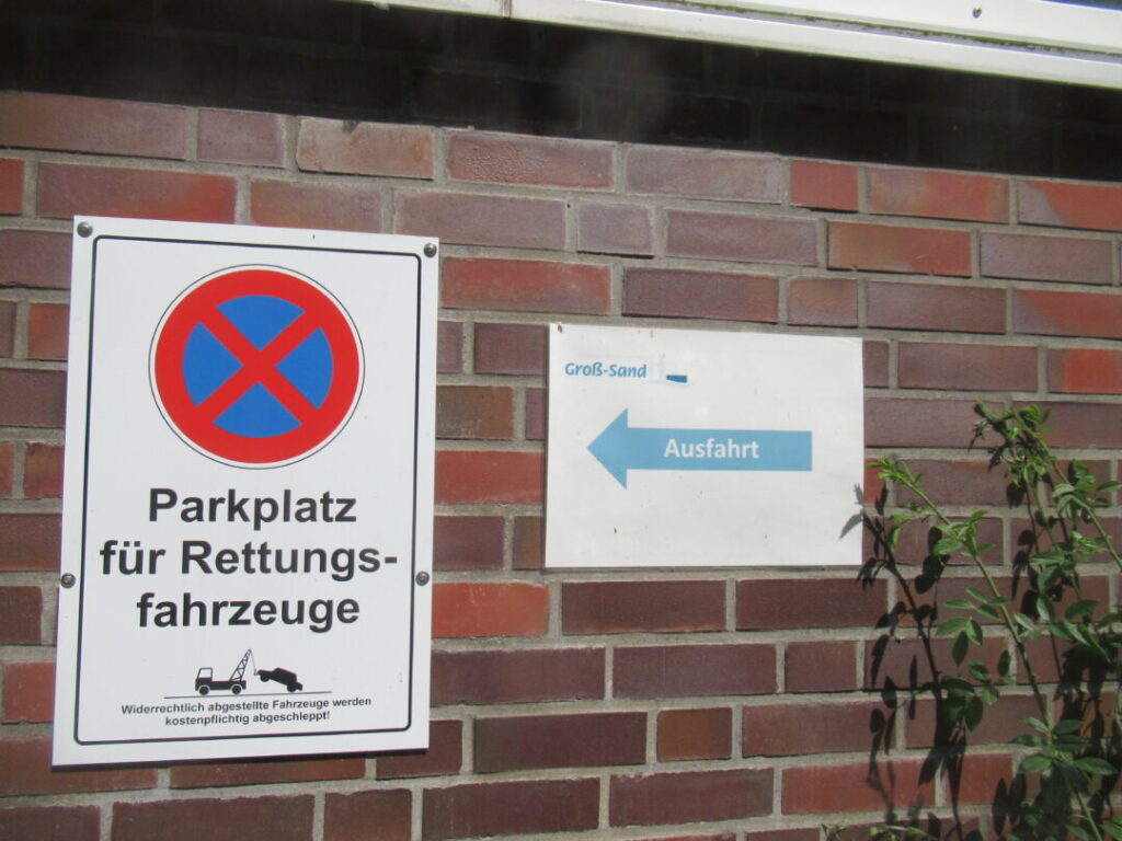 AN der Mauer das Halteverortsschild am Parkplatz für die Rettungsfahrzeuge, daneben ein Schild mit blauem Pfeil „Ausfahrt"