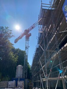 Links ein großer grüner Baum. In der Mitte steht ein hoher Kran und auf der rechten Seite ist ein Beton-Rohbau mit drei Stockwerken, davor ein Gerüst.