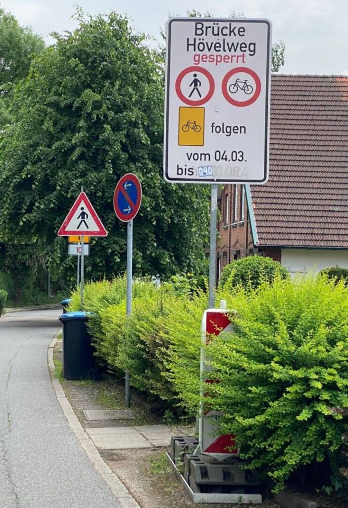 Die Schönenfelder Straße. rechts Büsche mit einer Papiertonne und drei Verkehrsschildern.