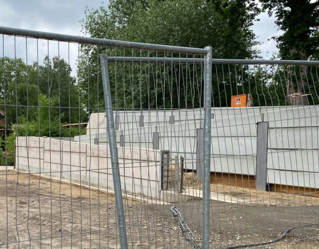 Vorne ein hoher Baustellenzaun und dahinter Betonmauern ca. 1 Meter hoch vor einem Baum.