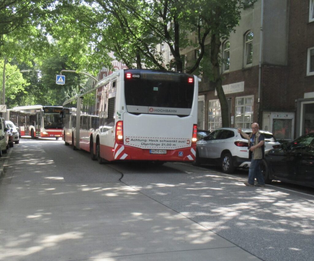 Im Vordergrund ein Mann, der einen Bus mit WInken zurücklotst. Im Hintergrund kommt der Gegenbus aus der Kurve.