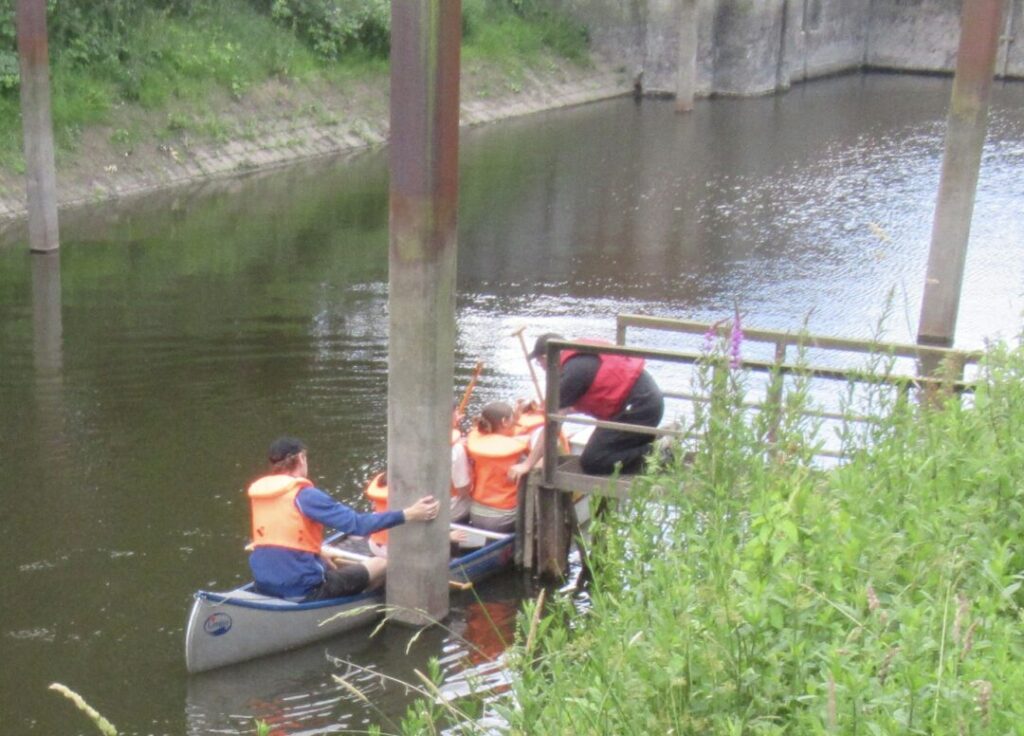Vier Kinder im Kanu legen an einer kleinen Anlegestelle am Ufer hinter der Schleuse an