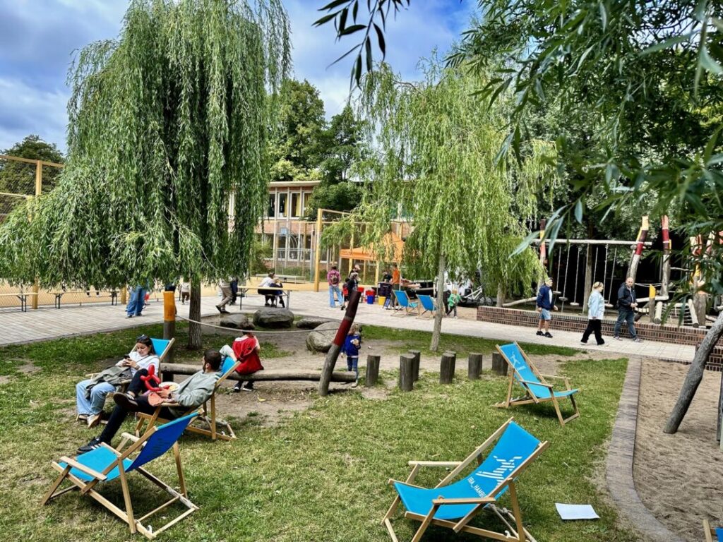 Auf einem großen Spielplatz in einer Grünanlage sitzen Menschen auf blauen Liegestühlen, im Hintergrund spielen Kinder.