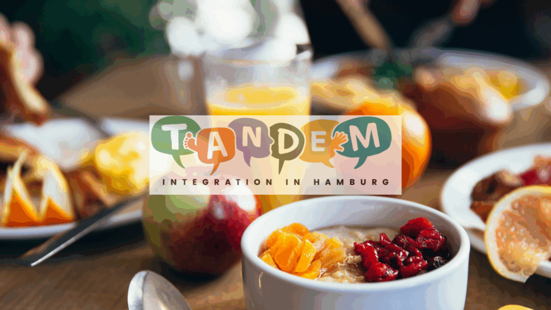 Ein gedeckter Frühstückstisch mit Müsli, Trockenfrüchten, frischem Obst, Muffins und einem Glas Orangensaft. In der Bildmitte ist das farbenfrohe Logo des Projekts "TANDEM – Integration in Hamburg" eingeblendet. Das Wort "TANDEM" ist in bunten Sprechblasen mit Symbolen wie Gesichtern, Händen und Füßen gestaltet, die für Kommunikation und kulturellen Austausch stehen.