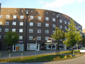 Ein rundes Wohnhaus auch Backsteinen, typisch für die Schumacherbauten auf der Veddel. Im Erdgeschoss Läden. Davor zwei kleine Bäume und ein freier Platz.