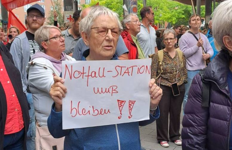 Demonstration. Im Vordergrund eine Teilnehmerin mit einem Schild: Notfallstation muss bleiben