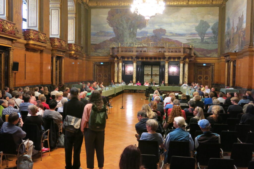 Blick in den Festsaal. Im Vordergrund die Besucher von hinten gesehen. Im Hintergrund die Ausschussmitglieder an U-förmig ausgerichteten Tischen mitn grünen Tischtüchern