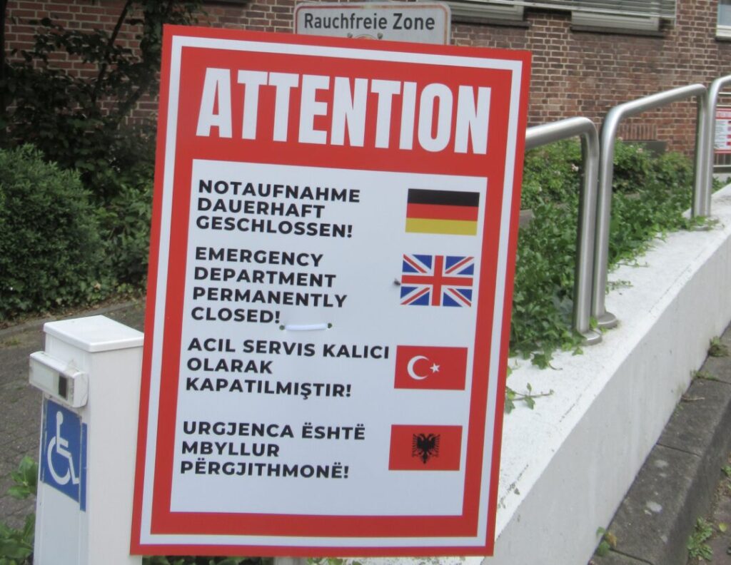 Schild auf dem Krankenhausgelände mit der Überschrft „Attention" und dem Text „Notaufnahme dauerhaft geschlossen" in vier Sprachen