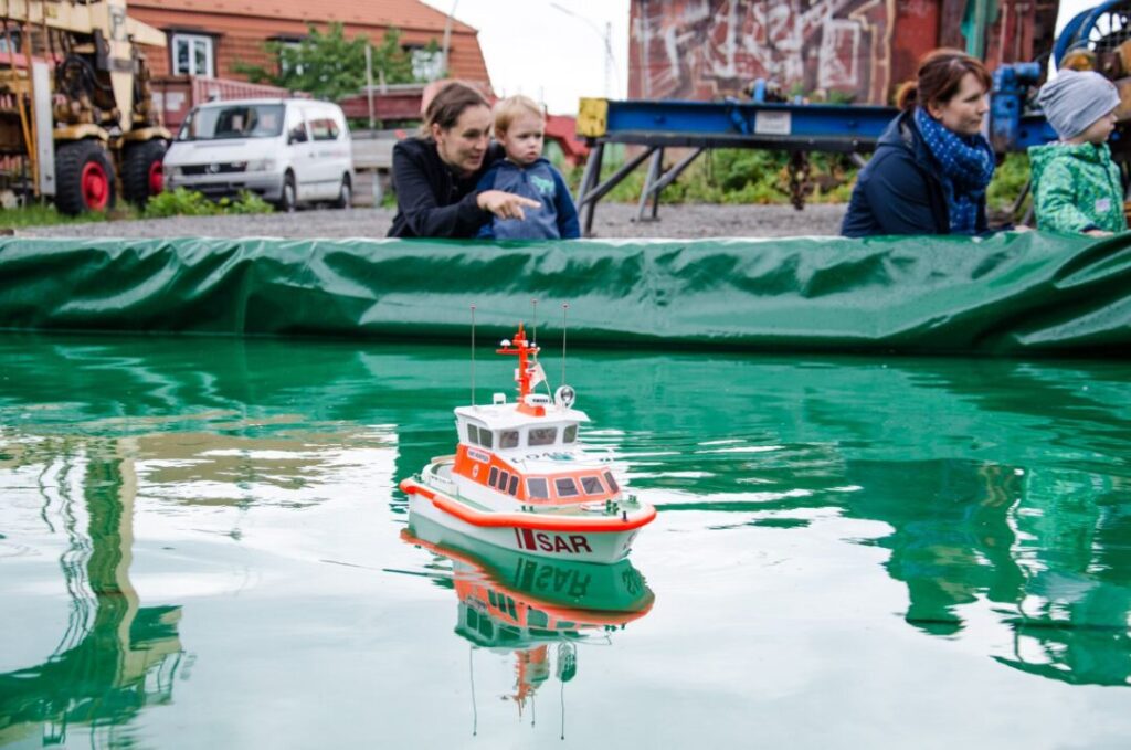 Im Vordergrund ein Wasserbecken aus grüner Plastikfolie mit einem Schiffsmodell. Am hinteren Rand zwei Mütter mit je einem Kind. Im Hintergrund Gebäude und ein Transporter.