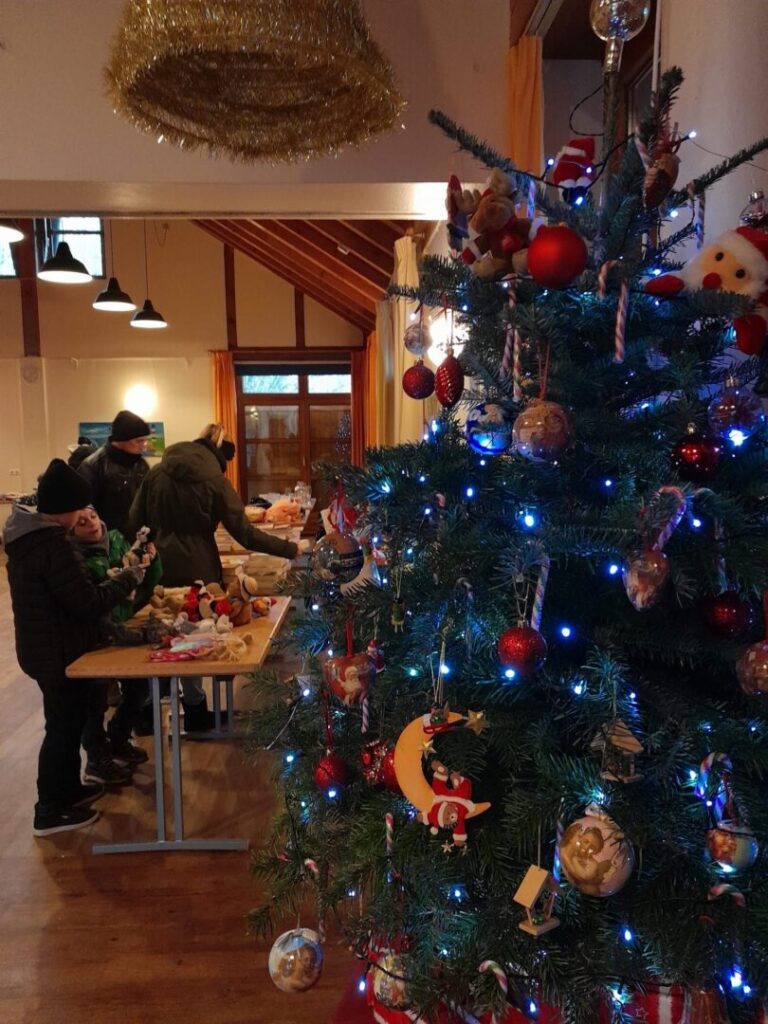 Im Freizeithaus zwei Menschen an Tischen mit Tauschgegenständen. Rechts im Bild ein bunt geschmückter Weihnachtsbaum.