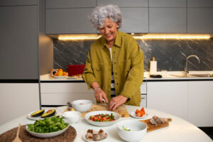 Eine ältere Frau mit grauen, lockigen Haaren steht lächelnd in einer modernen Küche und bereitet eine Mahlzeit zu. Sie trägt ein olivgrünes Hemd über einem gestreiften Oberteil. Auf dem runden Tisch vor ihr befinden sich verschiedene frische Zutaten wie Avocados, Erdbeeren, Pilze, Salat und Gewürze. Die Küche ist hell und stilvoll eingerichtet, mit grauen Schränken, Marmorrückwand und indirekter Beleuchtung unter den Oberschränken.