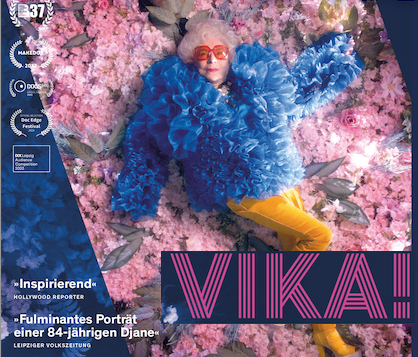 DJ Vika in blauer Jacke und gelber Hose räkelt sich auf rosa Blüten 