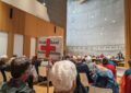 Menschen sitzen in einem hohen Raum. Links eine Holzwand und hinten eine Betonwand. Ein Schild mit einem roten Kreuz und Groß-Sand muss bleiben wird hoch gehalten.