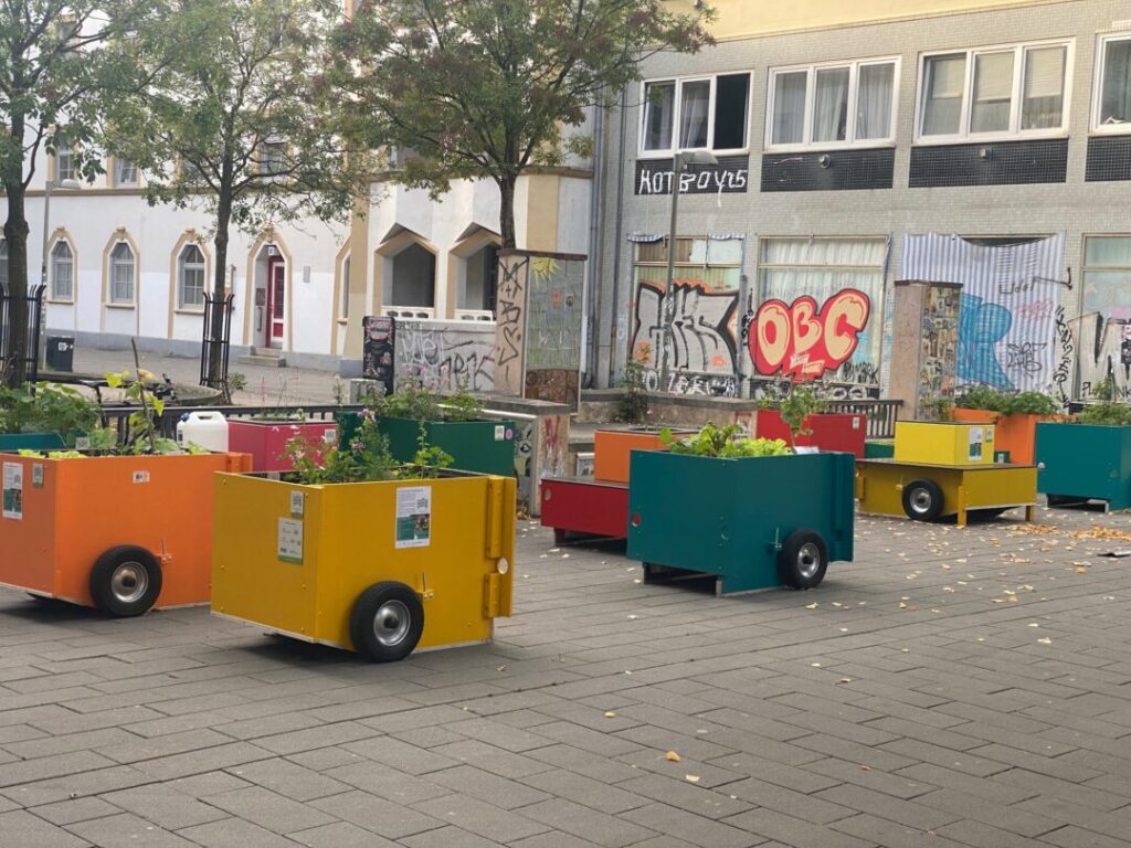 Kleine Hochbeete in Gelb, orange und blau mit einem Rad an der Seite stehen unter Bäumen auf einem Platz vor Jugendstilhäusern