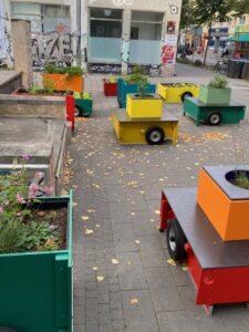 Auf grauen Platten stehen gelbe, orange, grüne, rote und blaue Kisten mit Rädern vor einem Haus mit Grafitto. Rechts oben sieht man die Veringstraße mit Bäumen.