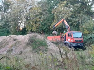 In der unteren Hälfte links ein Schuttberg. Daneben rechts ein roter LKW mit einem Kran. Im Vordergrund Wildwuchs und im Hintergrund Bäume und Büsche.