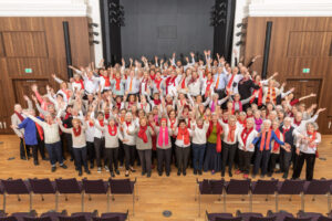 Hunderte Sängerinnen und Sänger in weißen Oberteilen mit roten Kravatten oder Schals vor einer Holzwand auf einer Bühne