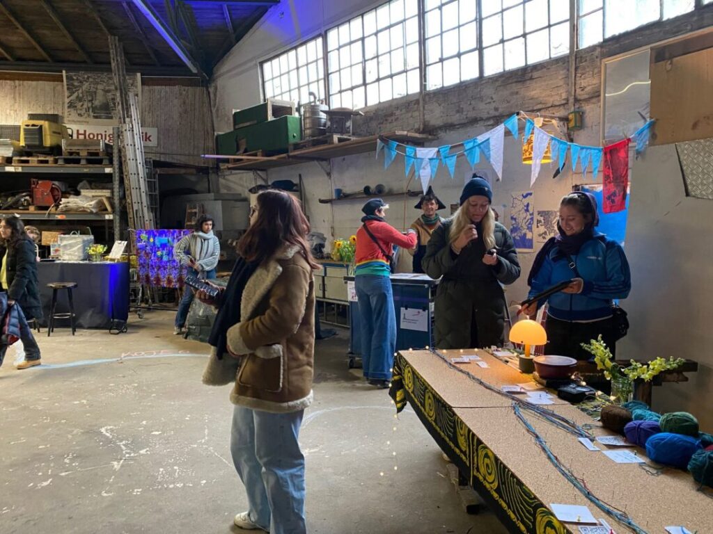 Im Vordergrund eine Frau. Rechts Installation mit Wolle. Dahinter eine Leine mit Dreiecken. Darüber eine Fensterfront. Im Hintergrund Menschen und ein Regal.