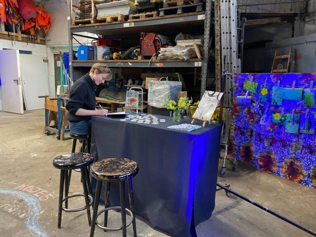 Im Hintergrund ein Materialregal. Davor eine Frau auf einem Barhocker an einem blauen Tresen. Davor zwei Barhocker.