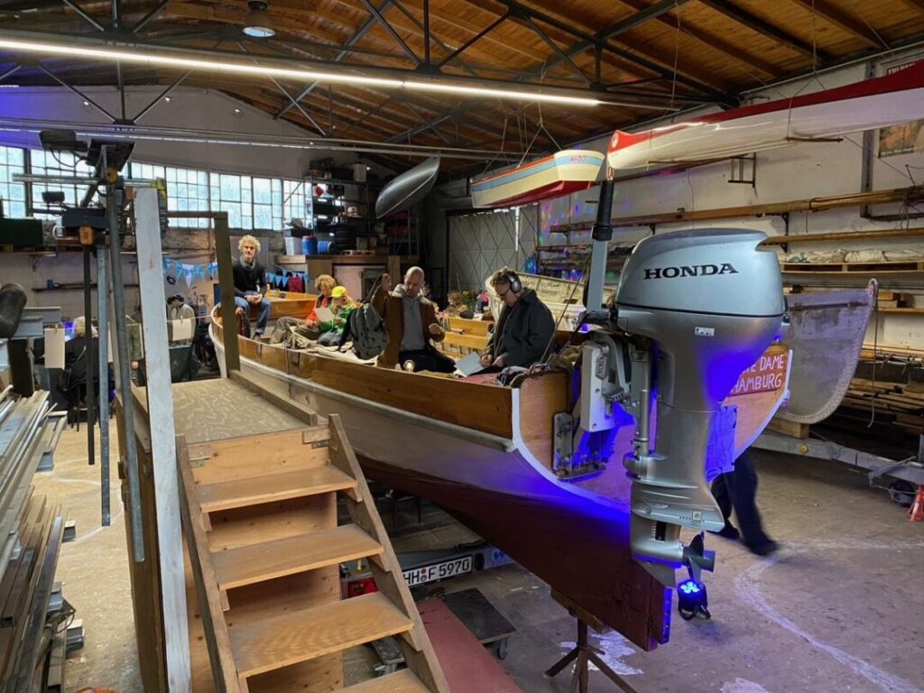 Boot in der Bootswerkstatt mit Menschen. Links daneben eine Treppe zu dem Boot. Hinten am Boot der Motor.