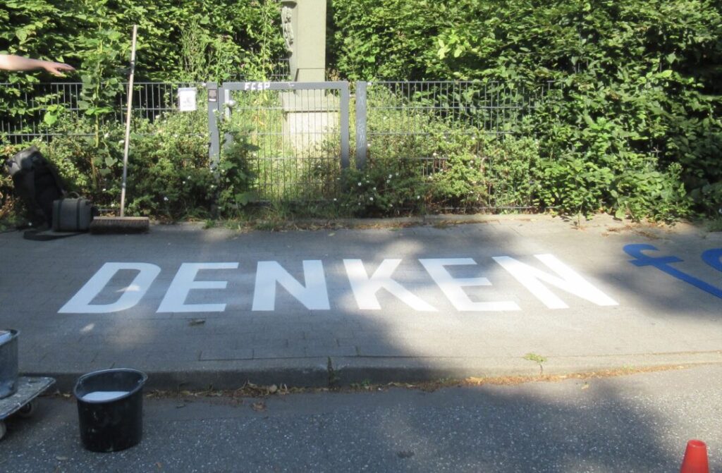 Auf dem Gehweg vor dem Denkmal ist in großen weißen Buchstaben das Wort DENKEN aufgebracht.