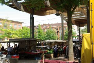 Wochenmarkt auf dem Stübenplatz unter dem gewellten Dach. Im Hintergrund die Häuser.