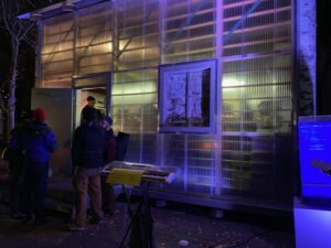 Im Dunkeln das Labor mit einer durchsichtigen Front, blau angeleuchtet. Links einige Menschen.