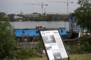 Im Hintergrund Kräne und Gebäude, davor Wasser und davor zwei blaue Havenlieger. Im Vordergrund vor einer kleinen Brücke ein Erklärungsschild.