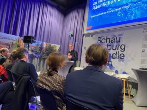 Mehrere Menschen sitzen im Vordergrund. Im Hintergrund ist ein blauer Vorhang, davor ein Mann mit Mikrofon an einem Stehtisch. Dahinter zwei Schautafeln, die obere blau, die untere weiß.