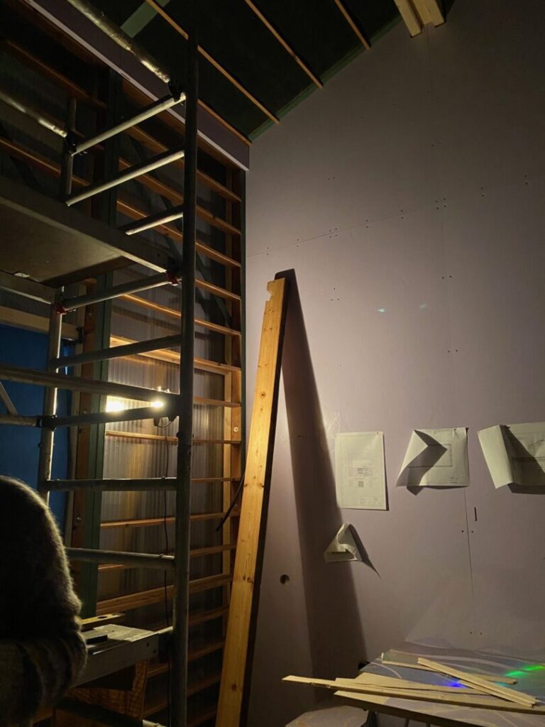 Rechte Hälfte eine beige Wand mit drei Zetteln. Ein Brett ist angelehnt. Linke Hälfte quer angebrachte Bretter. Wie ein Regal.