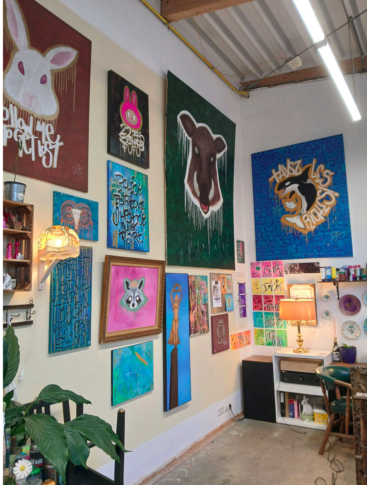 Eine Innenansicht eines Ateliers oder Kreativraums mit einer Wand, die dicht mit verschiedenen Kunstwerken in unterschiedlichen Stilen und Größen bedeckt ist. Links oben: Ein großes, dunkelrotes Bild mit einem weißen Hasenkopf. Mitte oben: Ein langes, vertikales Bild auf dunkelgrünem Hintergrund, das den Kopf eines Tieres (möglicherweise eine Tapir) zeigt. Rechts oben: Ein großes, blaues Bild mit einer schwarz-weißen Orca-artigen Figur und einem gelben Schriftzug. Darunter hängen kleinere, bunte Bilder in verschiedenen Formaten, darunter ein Bild mit einem Waschbären in einem rosa Rahmen, ein vertikales Bild einer stilisierten weiblichen Statue, und mehrere Bilder mit Text- oder Graffiti-Elementen auf türkisfarbenem und schwarzem Hintergrund. Rechts im Bild sind viele kleine, quadratische Kunstwerke aneinandergereiht. Links steht ein dunkler Stuhl und eine Pflanze. Eine kleine Holzkommode oder ein Regal ist mit verschiedenen Gegenständen wie kleinen Vasen und einem Eimer bestückt. In der Mitte leuchtet eine Tischlampe mit einem gewebten, gelben Lampenschirm und sorgt für eine warme Beleuchtung. Rechts steht ein Schreibtisch oder eine Ablagefläche mit einer weiteren Lampe und einem braunen Stuhl, umgeben von weiteren Dekorationsgegenständen und Farbdosen auf Regalen. Die Beleuchtung besteht aus Neonröhren an der Decke und warmen Tischlampen, was eine Mischung aus Arbeitslicht und gemütlicher Atmosphäre erzeugt. Der Boden ist Beton oder Estrich.