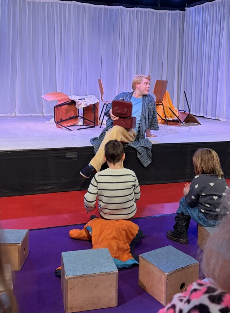 Eine Bühne, davor auf Hockern Kinder (Publikum). Am Bühnenrand sitzt der Hauptdarsteller in einem blauen Bademantel mit einer Kasse in der Hand. Hinter ihm liegen umgefallene Stühle.