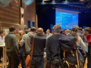 Rechts oben die Powerpoint-Präsentation. Links die Backsteinwand des Bürgerhauses. In der unteren Hälfte Menschen stehend dicht gedrängt.