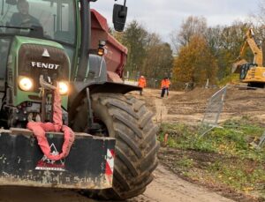 Eingrüner FENDT-Bagger im Vordergrund. Rechts eine Grünflähe mit einem Bauzaun. Im Hinergrund Bäume, davor zwei Menschen und rechts auf einem Sandhügel noch ein gelber Bagger.