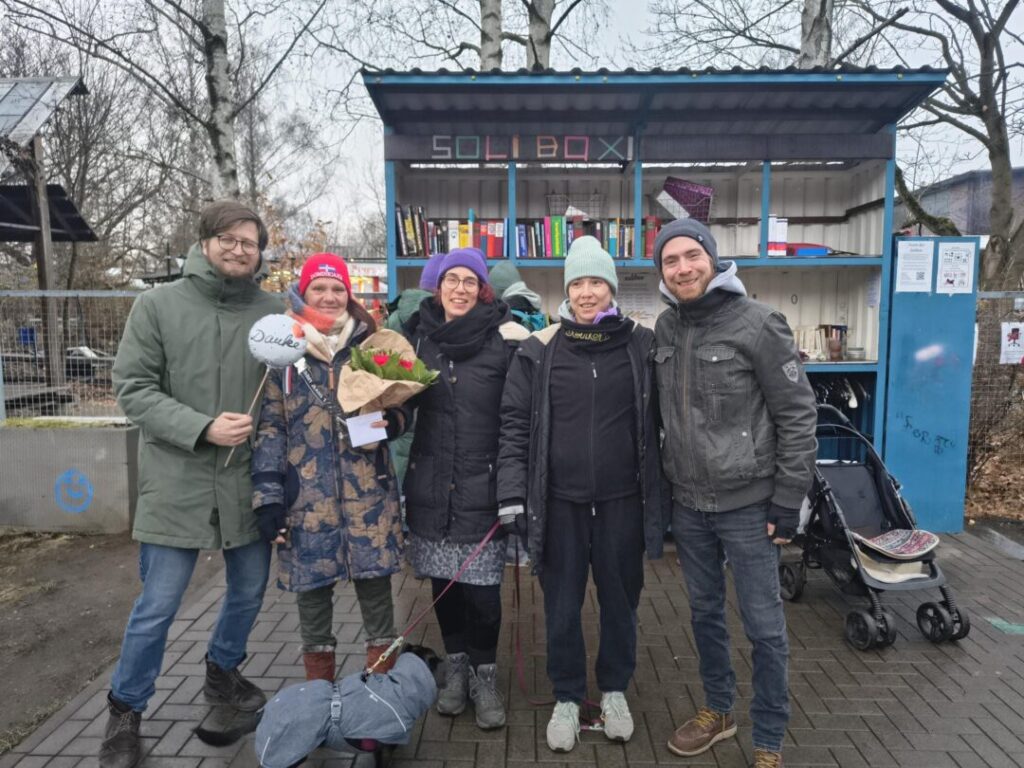 Das Orgateam, bestehend aus 5 winterlich dick angezogenen Personen steht vor der Soli-Box und strahlt in die Kamera.