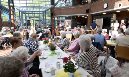 Im hellen Foyer des Bürgerhauses sitzen an langen Tischen viele ältere Menschen. Im Hintergrund ist zu erkennen, dass ein Künstler etwas sagt.