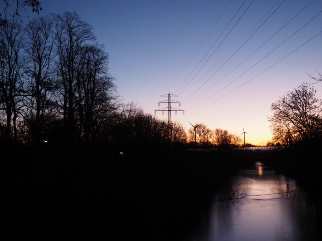 Blick vom Wilden Wald Richtung Osten: Zu sehen sind blattlose Bäume, eine Brücke über dem Kanal, eine Stromtrasse und zwei Windräder als Silhouetten vor dem langsam hellwerdenden Himmel. Foto: Angela Wolf