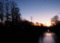 Blick vom Wilden Wald Richtung Osten: Zu sehen sind blattlose Bäume, eine Brücke über dem Kanal, eine Stromtrasse und zwei Windräder als Silhouetten vor dem langsam hellwerdenden Himmel. Foto: Angela Wolf