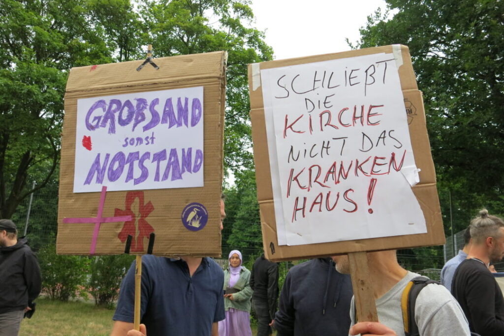 Zwei Demonstraneten, einer mit einem Pappschild: Schließt die Kirche, nicht das Krankenhaus