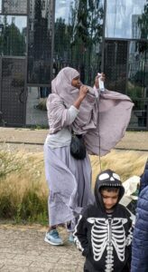 Eine Person, deren Kopftuch vom starkeWind verweht wird, spricht in ein Mikrofon. Im Hintergrund die Behörde, im Vordergrund ein Kind im Skelett-Anzug.