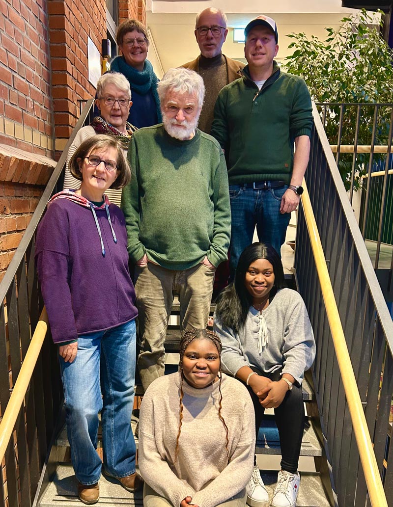 Ein Foto der Redaktion im Februar 2026: 8 Menschen auf einer Treppe. Im Hintergrund eine Backsteinmauer und ein Ficus Benjamini.
Foto: Melanie Brito