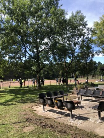 Sommer, hinter Bäumen ist ein Fußballplatz, am Rand stehen Zuschauende und gucken ein Spiel. Im Vordergrund sind Sitzplätze. Foto: Calle Stentzler