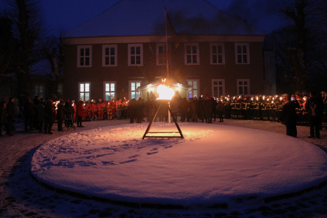 Vor einem großen Gebäude mit weißen Fenstern stehen in der Dunkelheit knapp 100 Menschen mit Fackeln. Davor in der Mitte auf einem Schneefeld eine große Fackel auf einem dreibeiniten Ständer