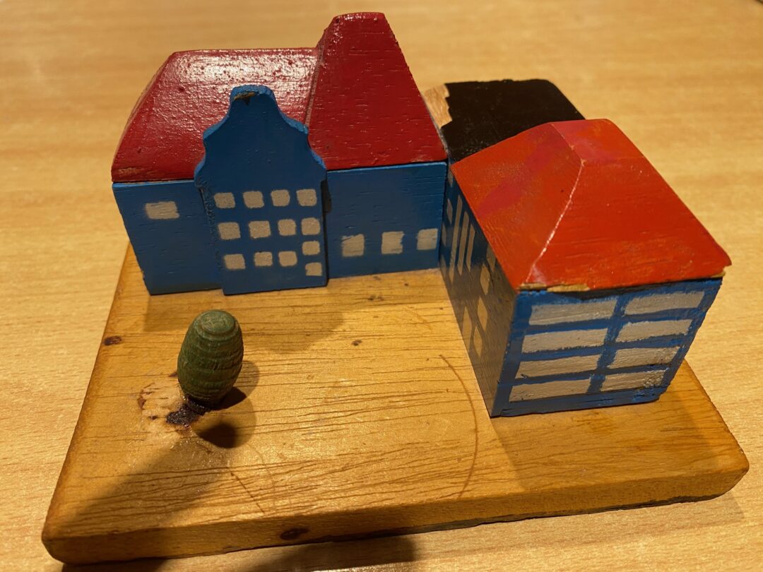 Auf einem hellen Holztisch liegt eine Holzplatte. darauf ein Holzmodell, hinten mit einem Erker in der Mitte. Rechts im 90 Grad-Winkel ein vierstöckiges Gebäude. Beide mit Fenstern und blau gestrichen mit einem leutend roten Dach.