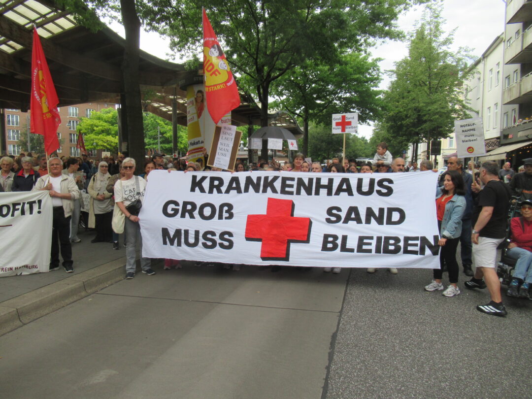 Kopf einer Demnstration mit ddem großen Transparent mit der Aufschrift: Krankenhaus Groß Sand muss bleiben und einem roten Kreuz in der Mitte