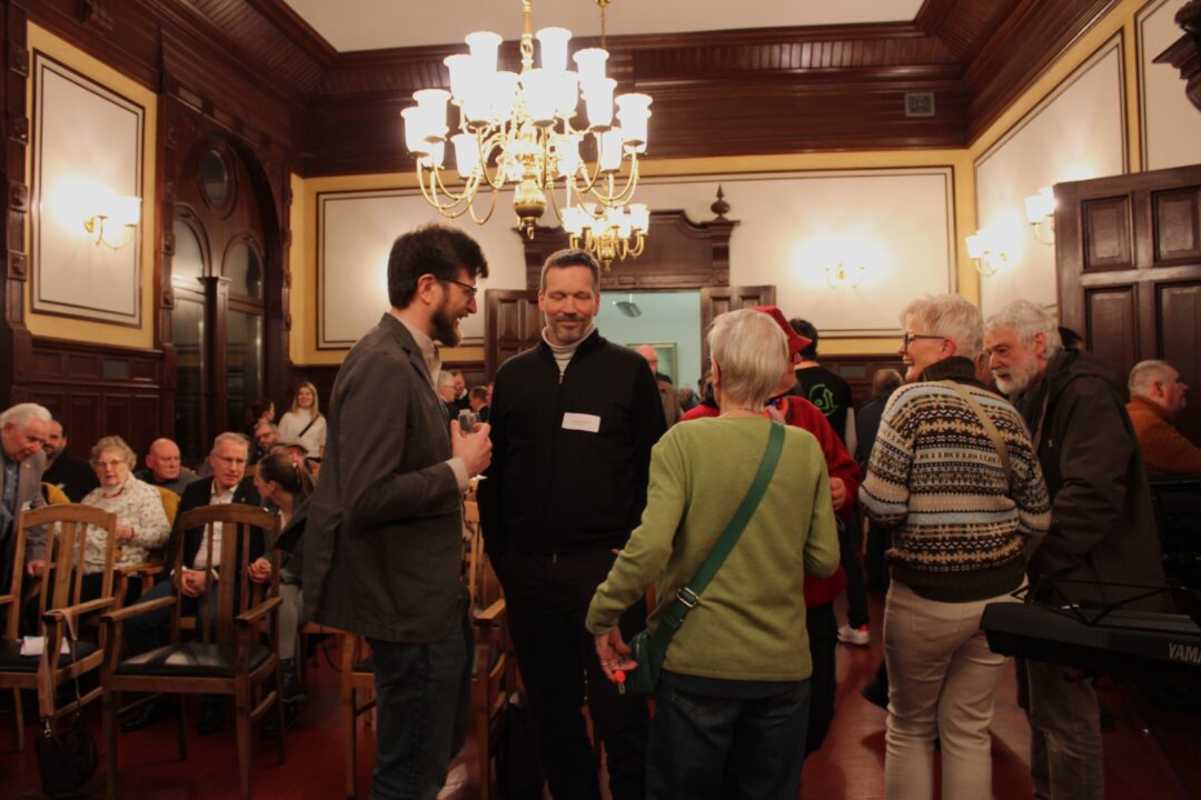 In einem historischen Sall mit Bildern an den Wänden sitzen mehrere Menschen. Im vordergrund stehen sechs Menschen ins Gespräch vertieft.