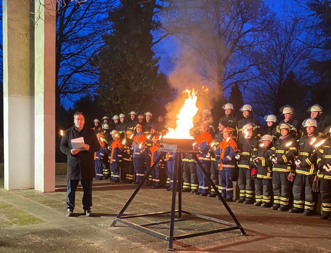 Links eine weiße Säule, davor ein Mann mit einem Manuskript in der Hand. Im Mittelpunkt ein vierbeiniges Gestell, oben die Feuerschale mit einer lodernden Flamme. Dahinter Menschen der Feuerwehr und Deichwacht.